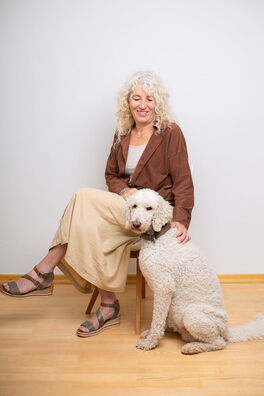 Petra Endres von Dialogwege sitzt auf einem Stuhl. Davor sitzt ihr Hund